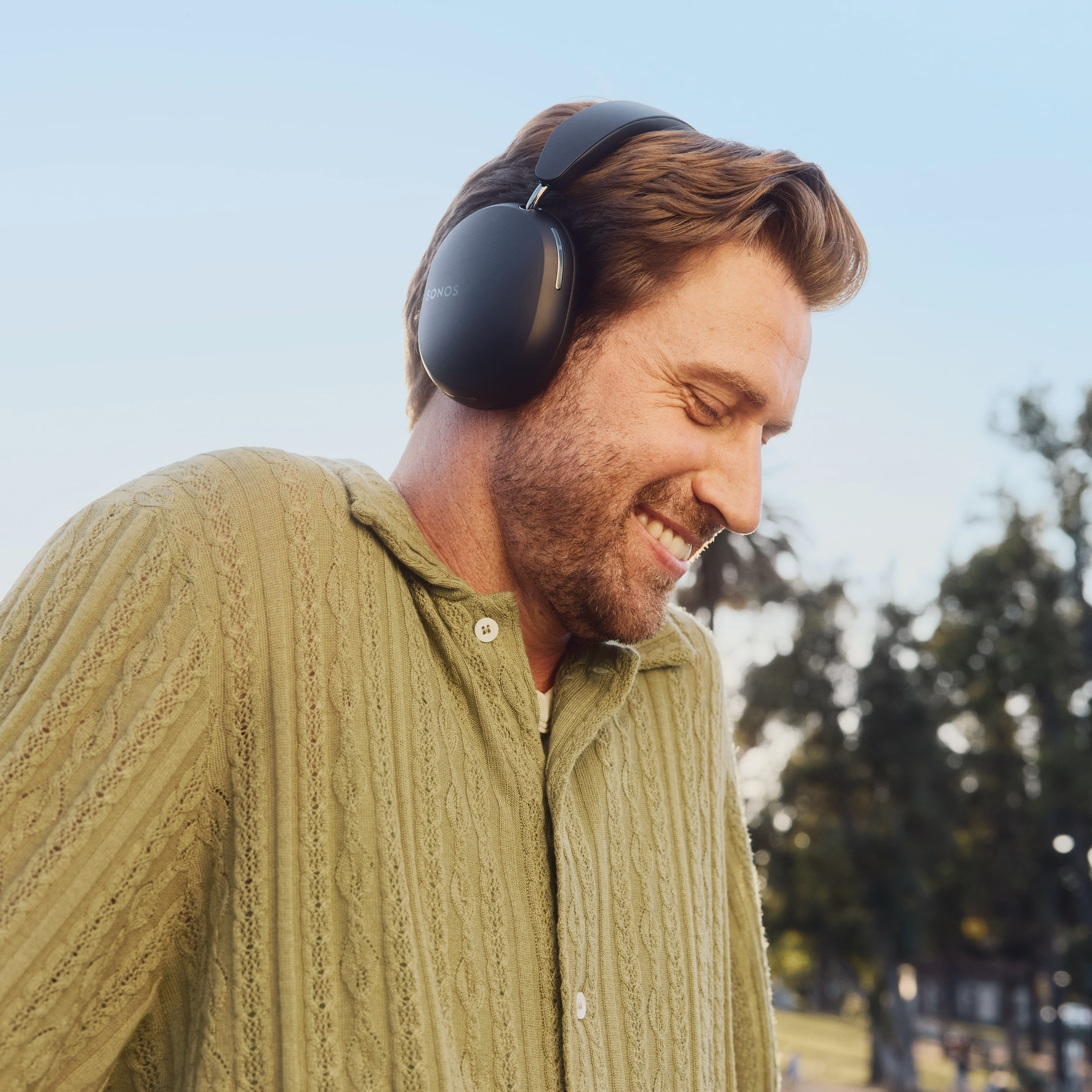 Person smiling listening to music on black Sonos Ace headphones in the park